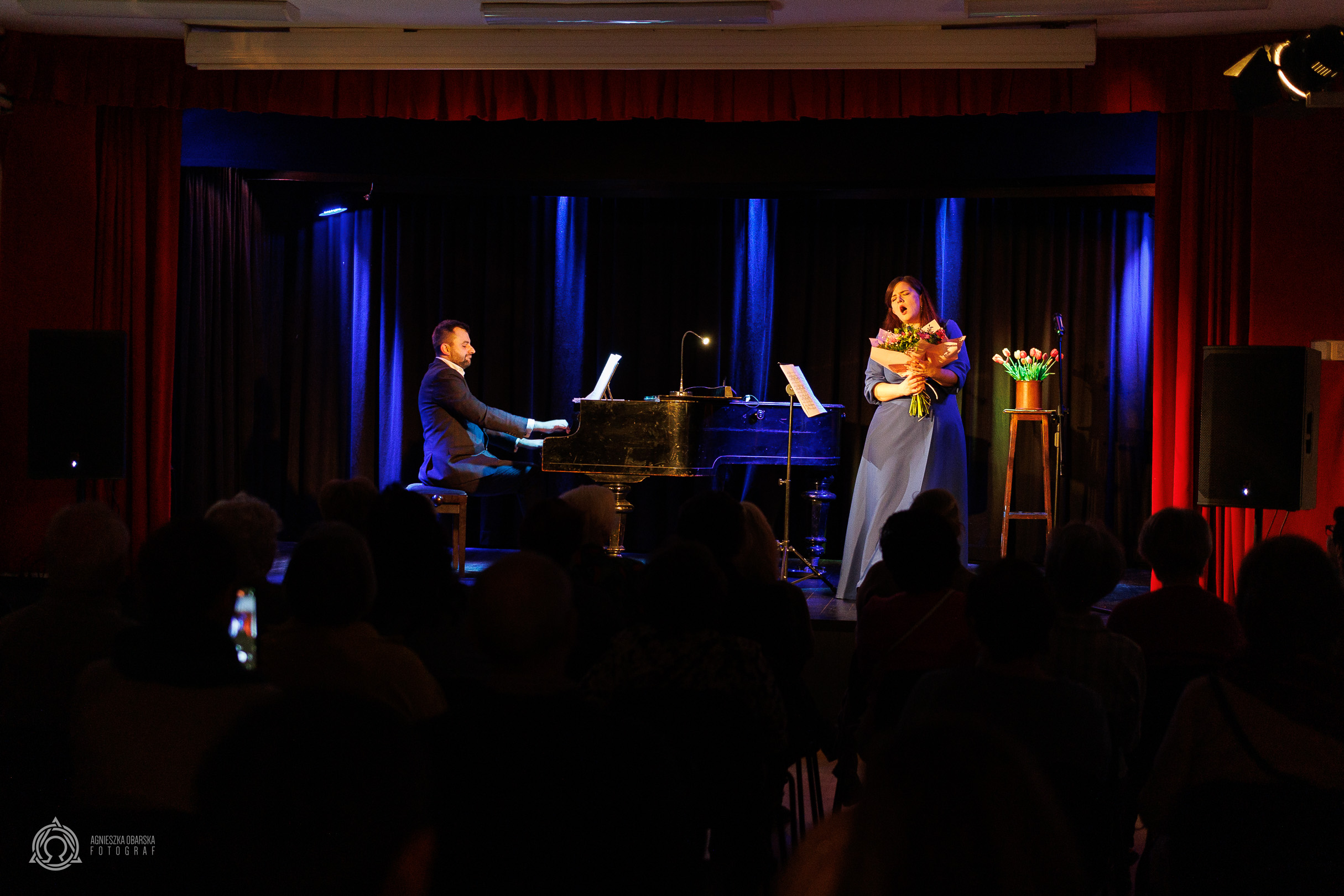 Ubrana w niebieską sukienkę kobieta śpiewa na scenie do muzyki granej na fortepianie, przez ubranego w garnitur mężczyznę.